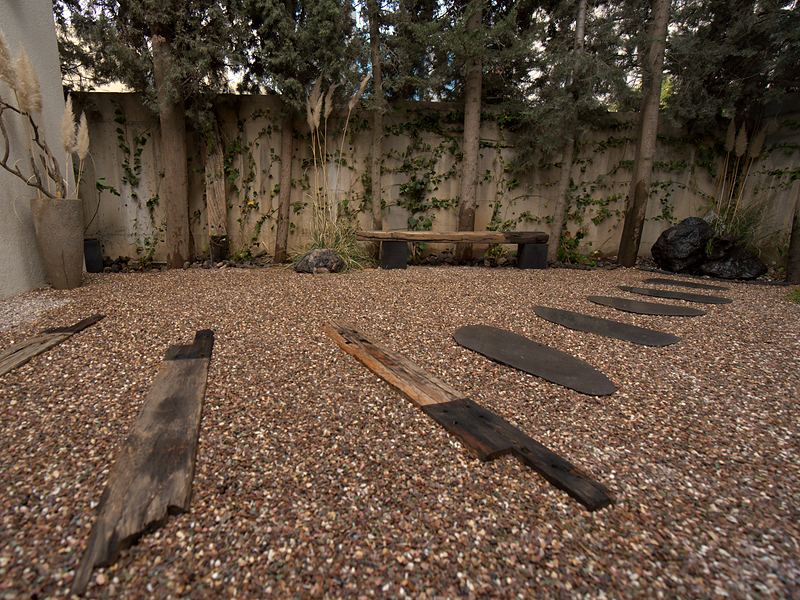A low view across a gravel courtyard: weathered timber planks and dark basalt stepping stones lead to a beam bench, with pampas grass, cypress trees and a vine-covered wall behind.